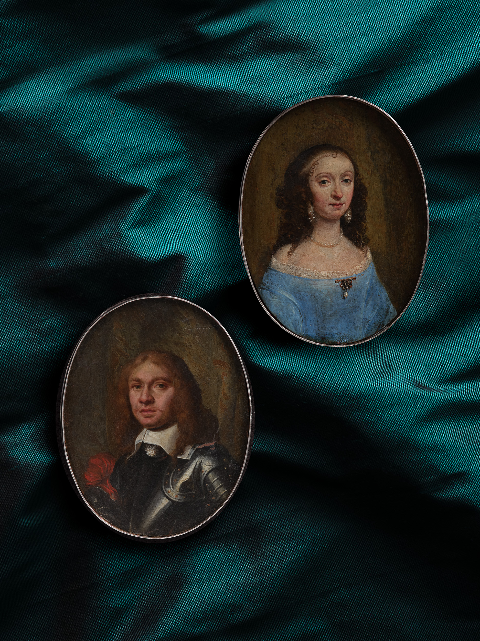 A pair of oil portraits, presumably Husband and Wife; he wearing armour, lawn collar and a red sash around his arm; she wearing lace-trimmed blue dress with diamond and pearl brooch, pearls and pearl-drop earrings