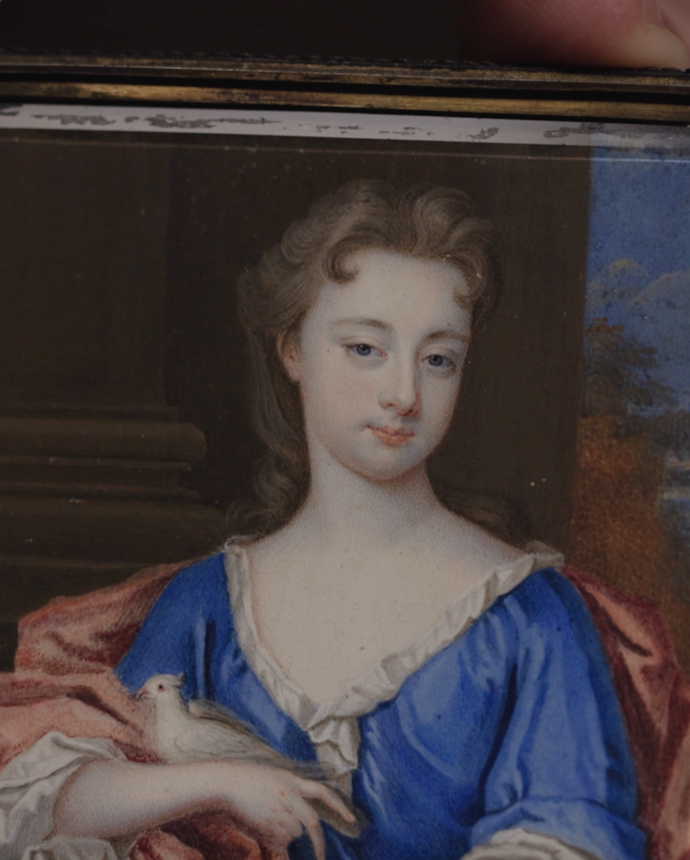 Portrait miniature of a Lady, seated in front of a column with landscape behind, wearing blue sink dress with white sleeves and collar, red cloak, holding a dove in her right arm