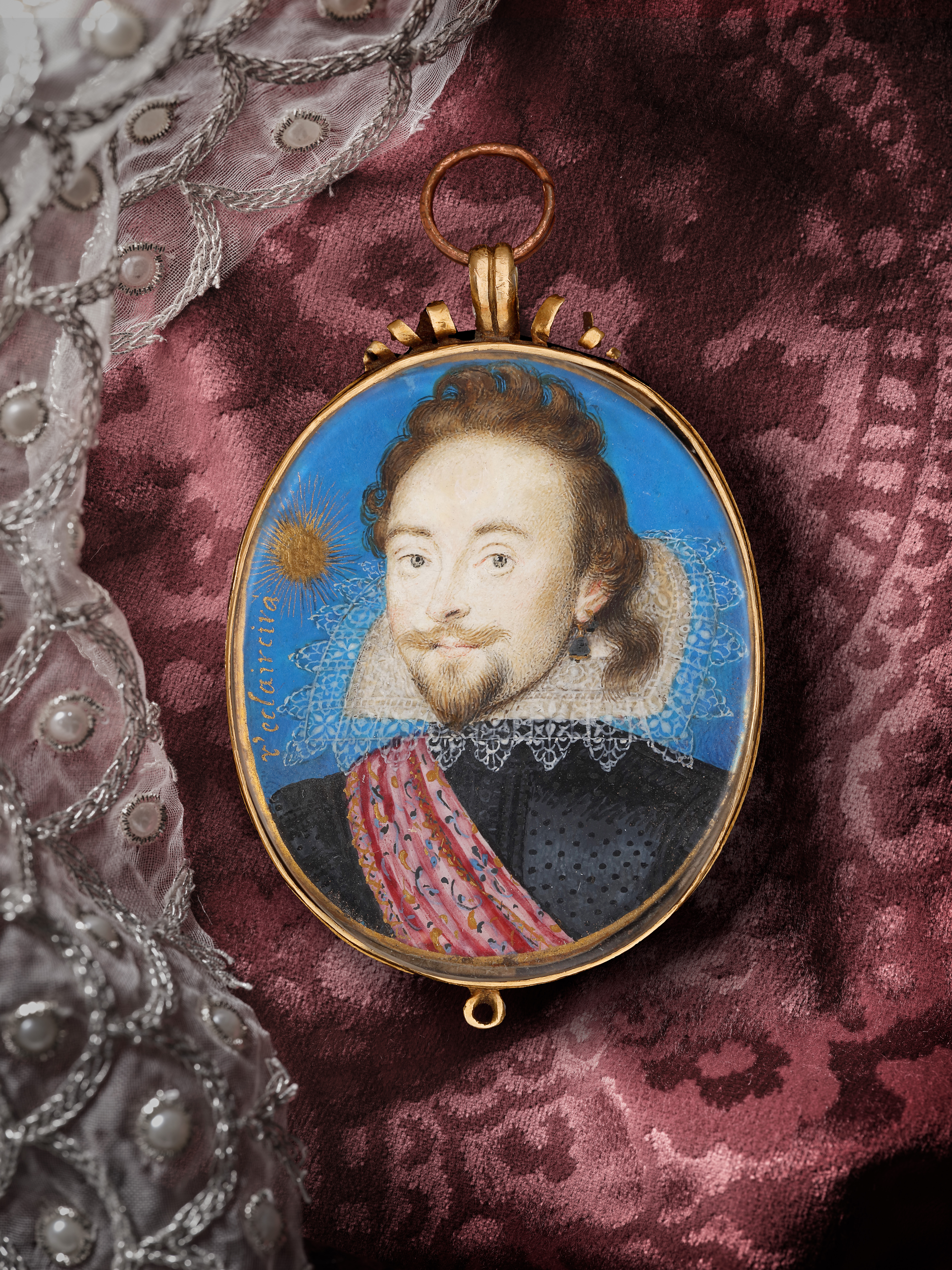 Portrait miniature of a Nobleman wearing a black doublet with an embroidered pink sash, a rebato collar with scalloped lace edging, and a large gem stone earring; his hair in a ‘lovelock’ on his left; a gold sunburst over his right shoulder